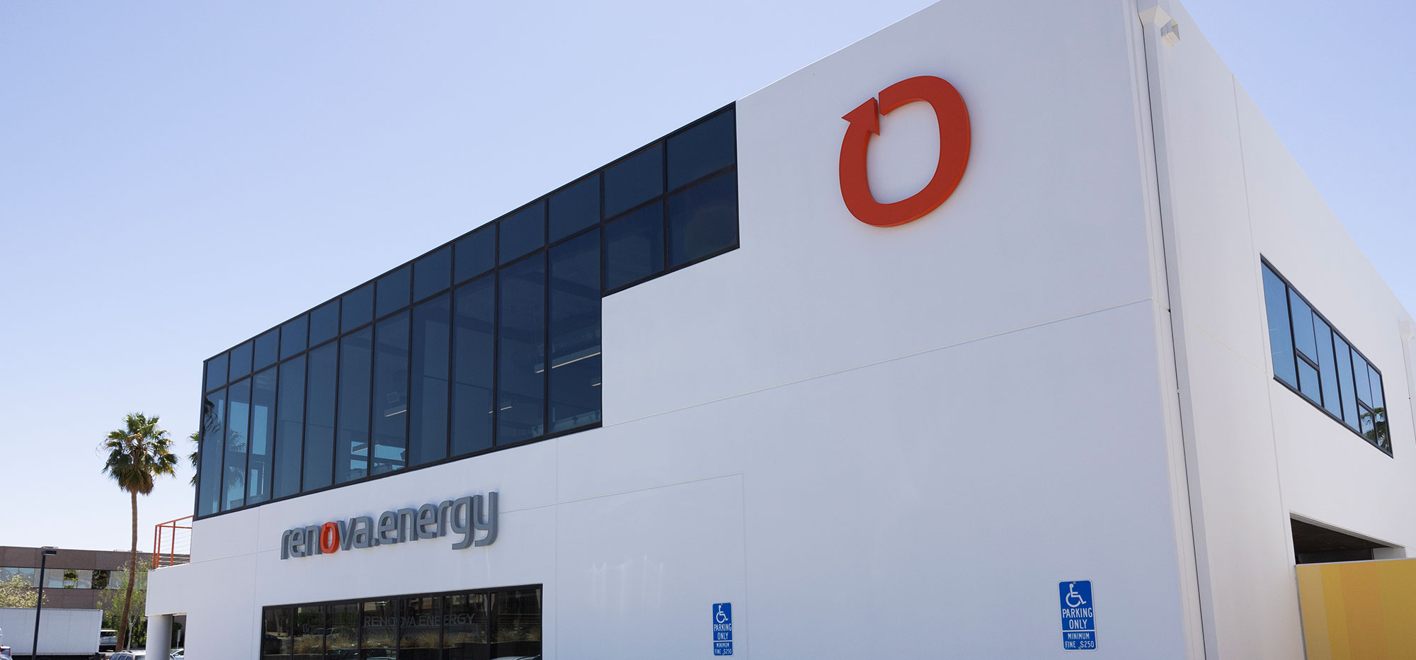 Modern white building with large dark windows and an orange circular logo on the upper corner. The words renewable energy are displayed on the side. A palm tree is visible to the left, and two handicapped parking signs are in front. Clear blue sky in the background.