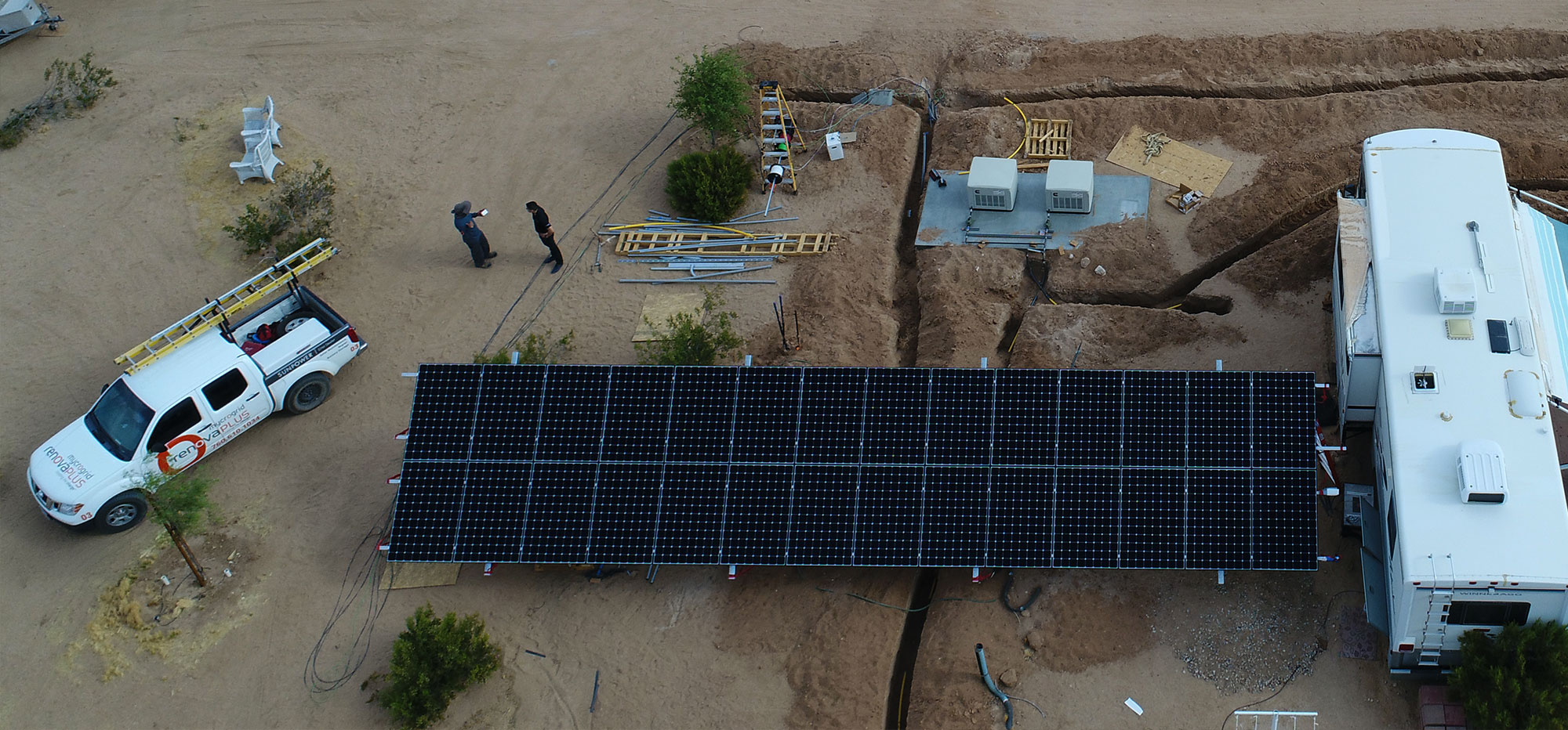 Aerial view of a desert scene with a large solar panel array connected to a white vehicle. Two people stand near a parked truck equipped with ladders. Nearby, there are scattered tools and equipment, with desert plants surrounding the setup.