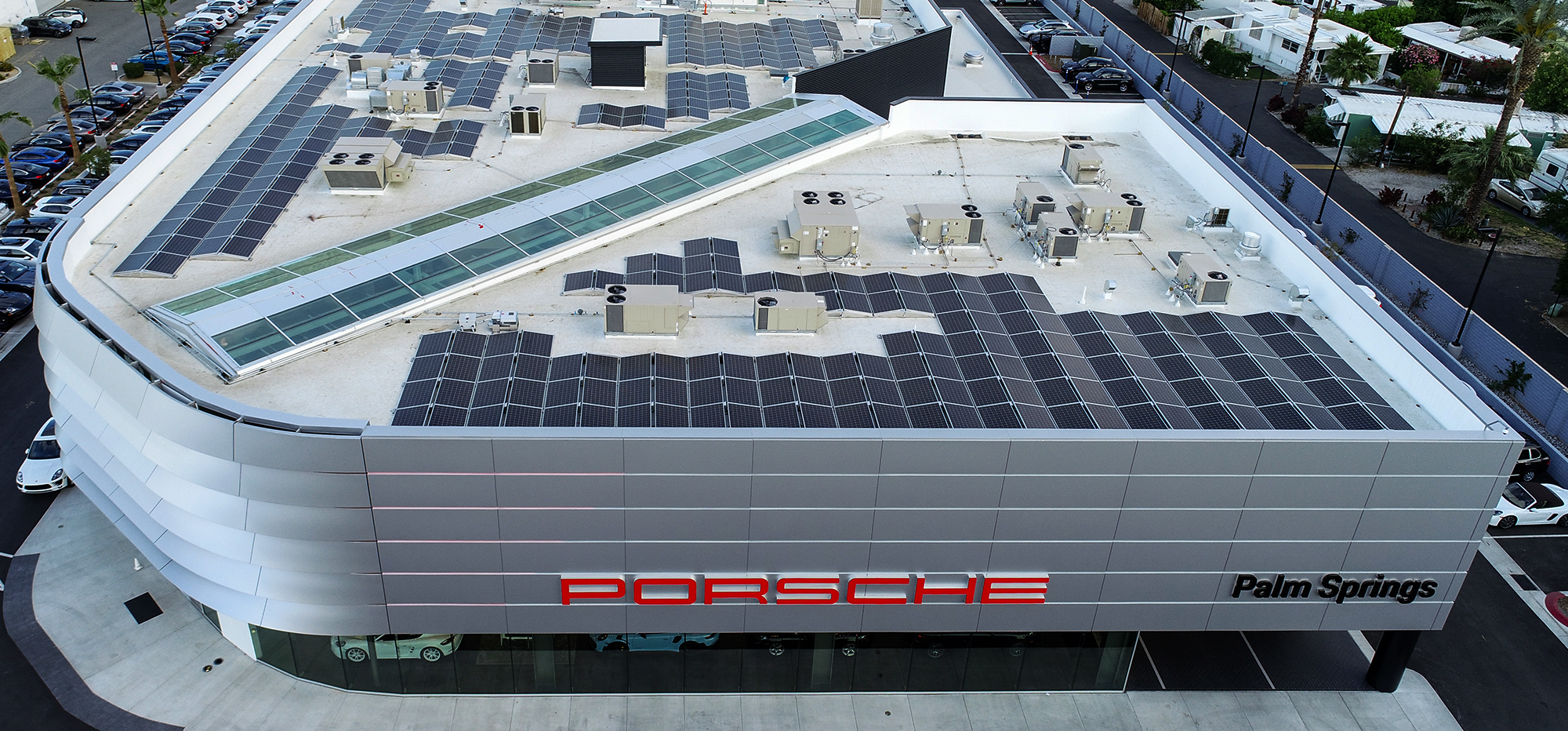 Aerial view of a modern building with Porsche in large red letters and Palm Springs in black on the facade. The rooftop is covered with solar panels and various HVAC units. The structure is surrounded by parked cars and greenery, suggesting a dealership location.