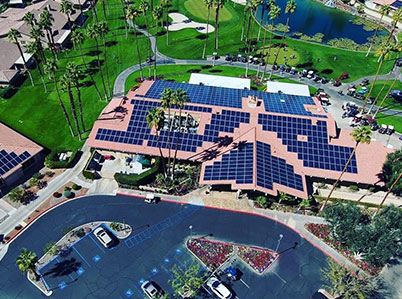 Aerial view of a building with solar panels on the roof, surrounded by green lawns, palm trees, and a golf course. Several parked cars are visible near the building. The setting appears to be a sunny day, highlighting the clean and energy-efficient environment.