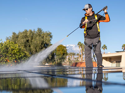 A person wearing a cap and sunglasses cleans solar panels with a water hose on a sunny day. Theyre equipped with a safety harness and surrounded by trees and palm trees, with a background of clear blue sky and distant mountains. The panels reflect the clear sky.