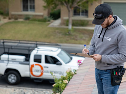 A person wearing a gray hoodie and cap, both with a logo, stands on a rooftop inspecting a clipboard with a pen. A white truck with a logo and roof rack is parked on the street nearby. Residential houses and a yard are visible in the background.