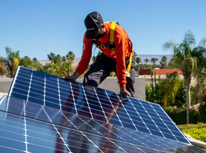 A worker wearing an orange safety harness and cap installs solar panels on a rooftop. The sky is clear, and palm trees are visible in the background.