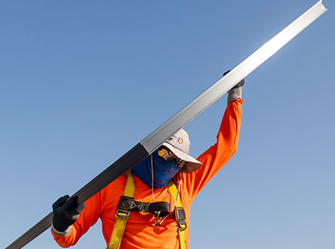 A construction worker in an orange shirt and safety harness holds a long metal beam. The worker is wearing a wide-brimmed hat, face covering, and sunglasses. The sky is clear and blue, creating a bright backdrop.