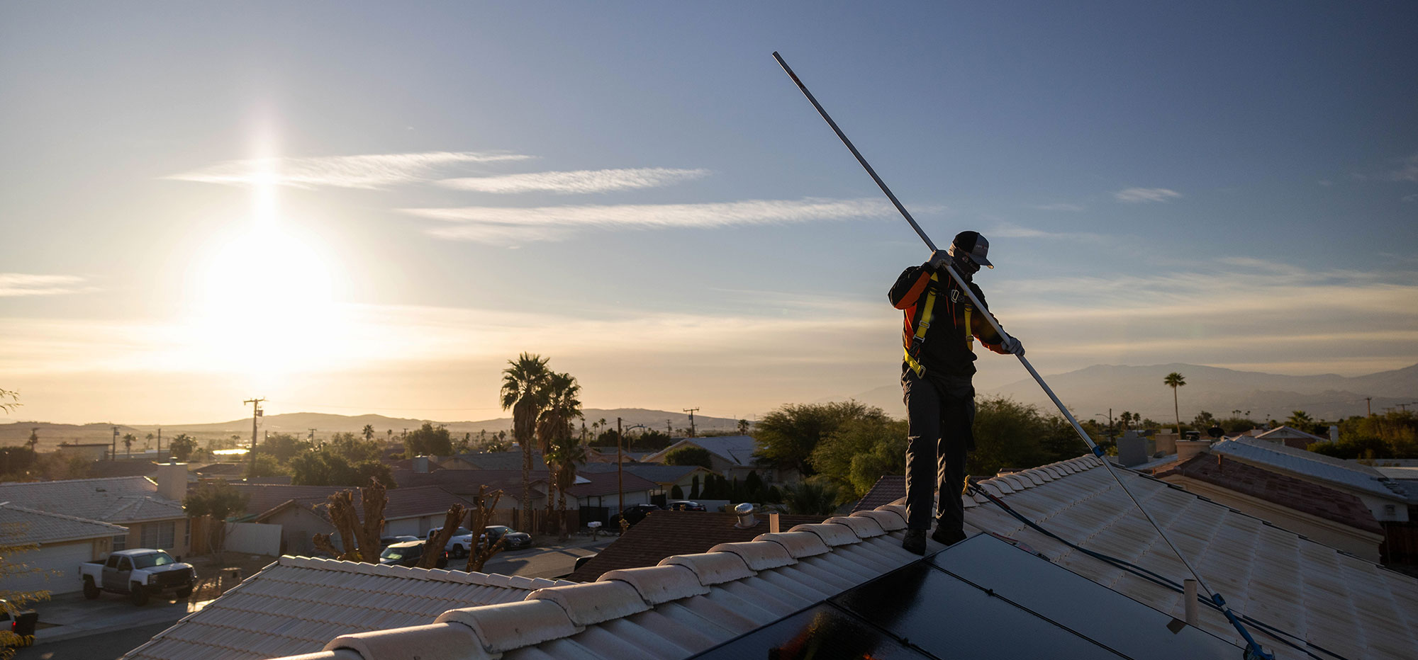 A person is cleaning solar panels on a rooftop at sunrise or sunset, using a long pole. They are wearing a harness for safety. The background features a suburban neighborhood with palm trees and mountains under a clear sky.