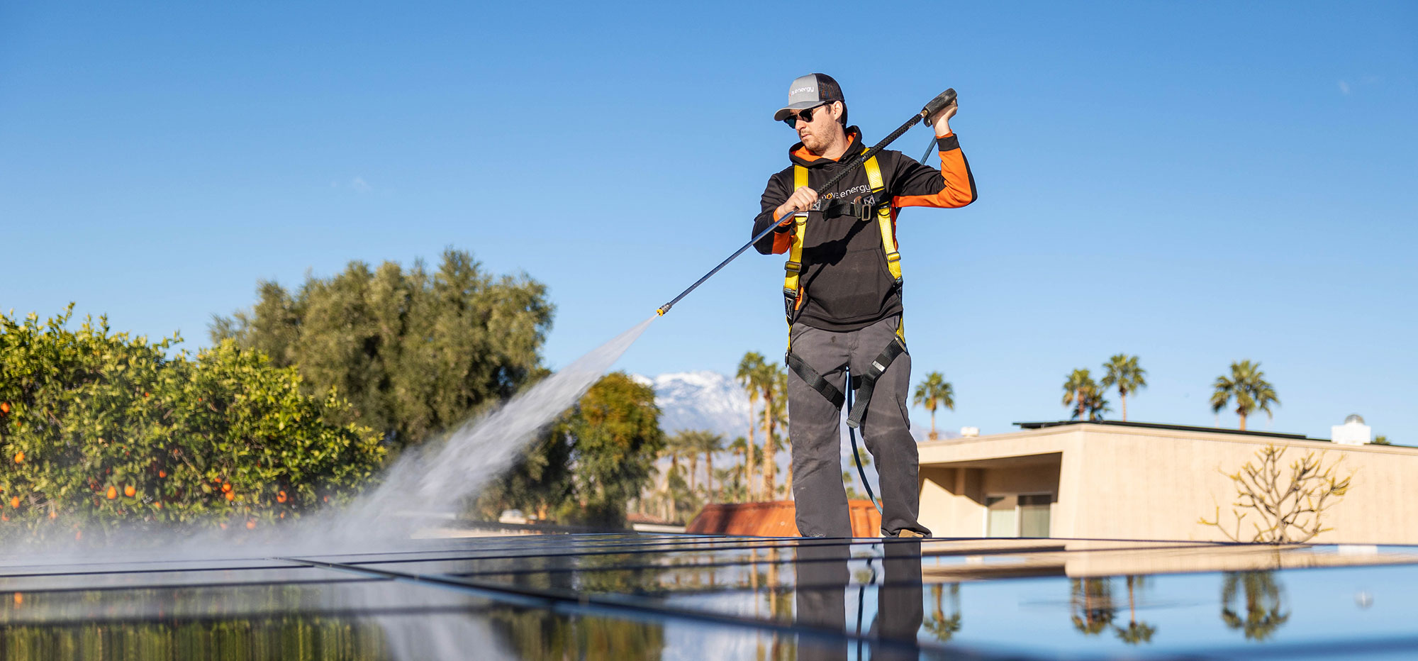 A worker wearing a cap, safety harness, and sunglasses uses a pressure washer to clean solar panels on a roof. The sky is clear and blue, with mountains and palm trees in the background. The worker is focused on his task, and water sprays across the panels.