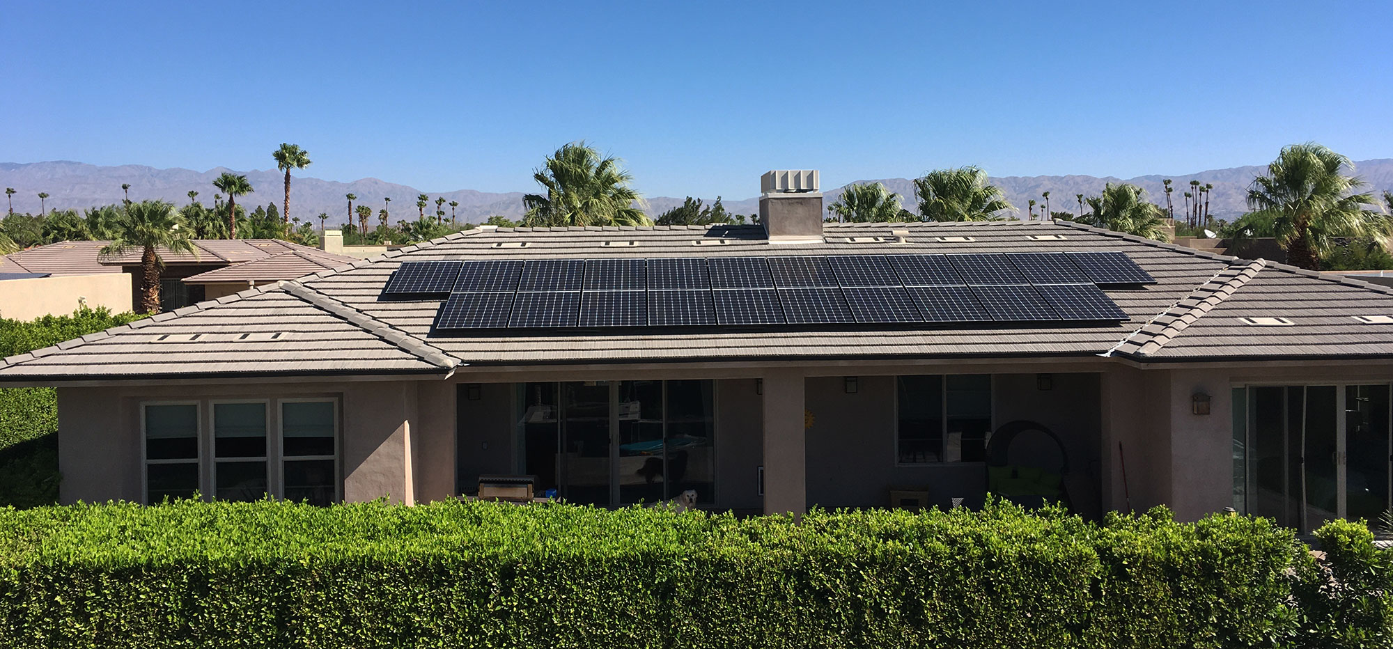 A single-story beige house with a tile roof features multiple solar panels. Lush green hedges surround the property, and tall palm trees are visible in the background. Beyond the trees, distant mountains can be seen under a clear blue sky.