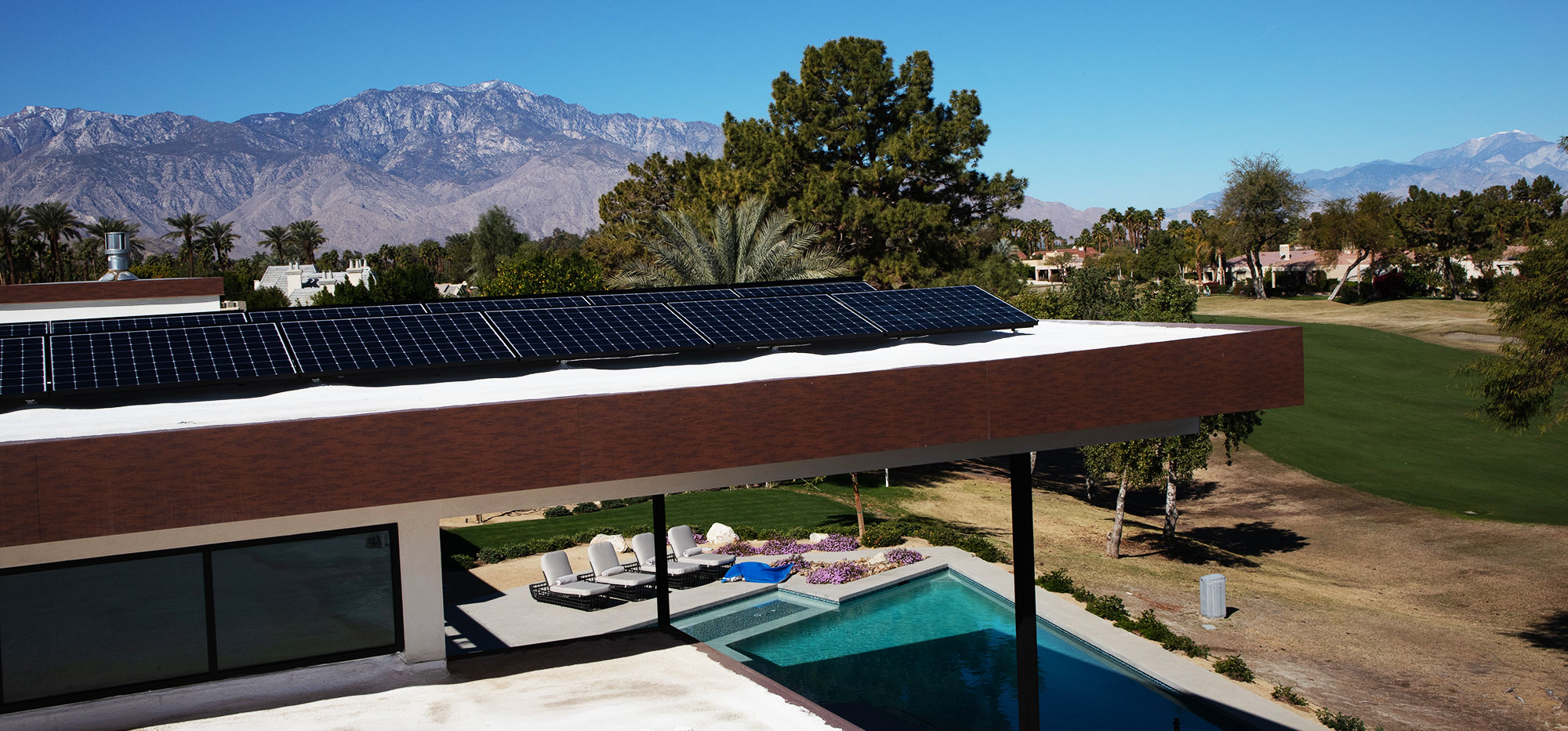 A modern house with a flat roof fitted with solar panels overlooks a landscaped golf course. A blue swimming pool sits adjacent to the house, surrounded by lounge chairs. Mountains and a clear blue sky are visible in the background, creating a serene setting.