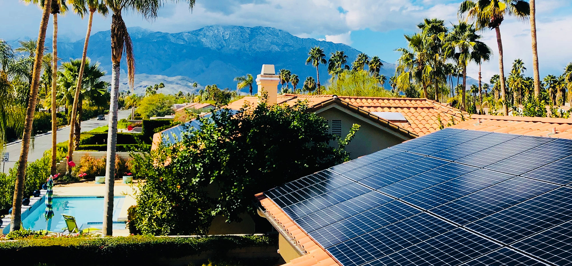 A rooftop with solar panels is in the foreground, surrounded by lush greenery and palm trees. Behind the house, a clear swimming pool is visible. In the distance, tall mountains are under a partly cloudy sky, suggesting a sunny, warm climate. A quiet road lines the side of the property.