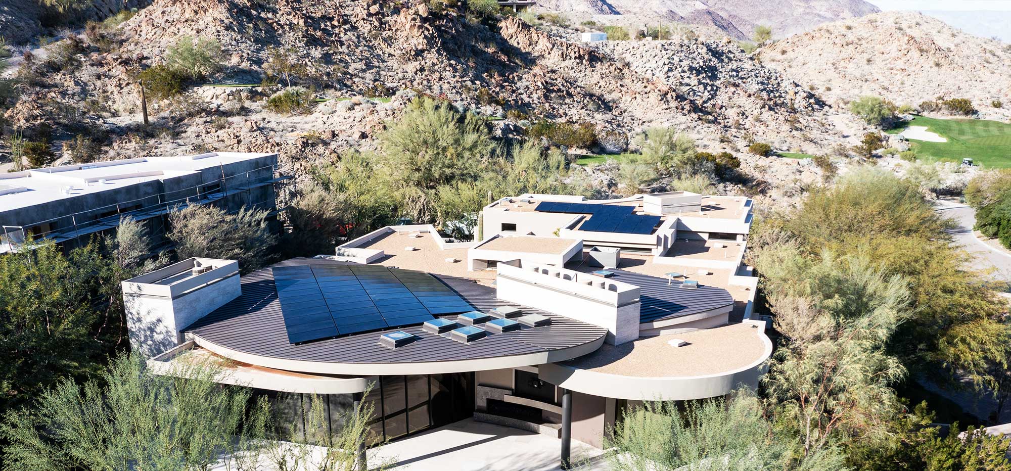 A modern hillside home with a flat roof, solar panels, and circular architectural elements nestled among rocks and desert vegetation. The house features large windows and several terraces, set against a backdrop of mountains. Greenery surrounds the structure, blending with the natural landscape.