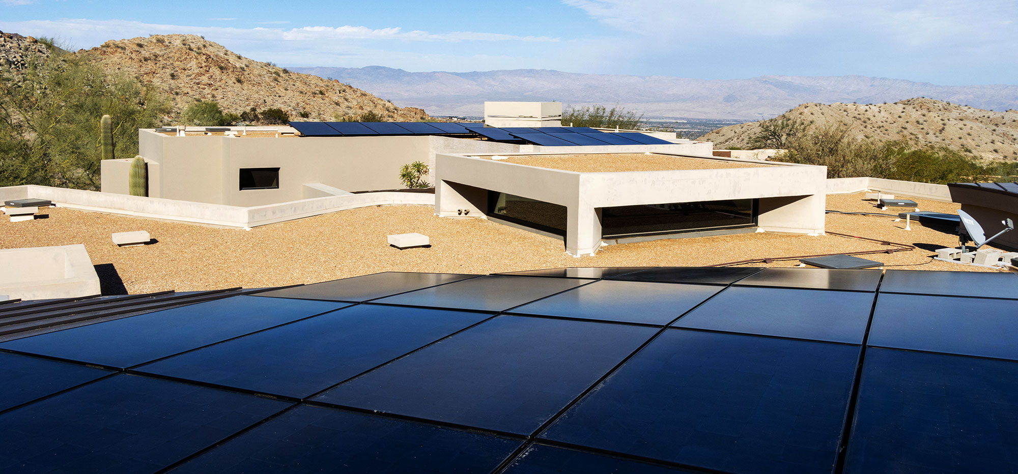 A modern house with a flat, gravel-covered roof features multiple solar panels. The surrounding desert landscape includes mountains and scattered vegetation under a clear blue sky. The architecture is minimalist with geometric shapes, and the scene conveys a sense of sustainable living.