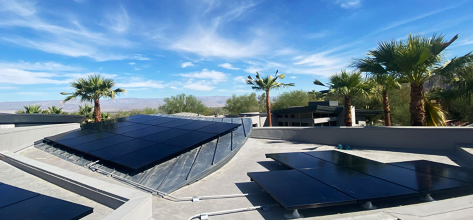 A rooftop with multiple solar panels under a bright blue sky, scattered with wispy clouds. Surrounding the roof are tall palm trees and distant hills, suggesting a desert-like environment. The scene conveys a sense of renewable energy in a sunny, arid location.