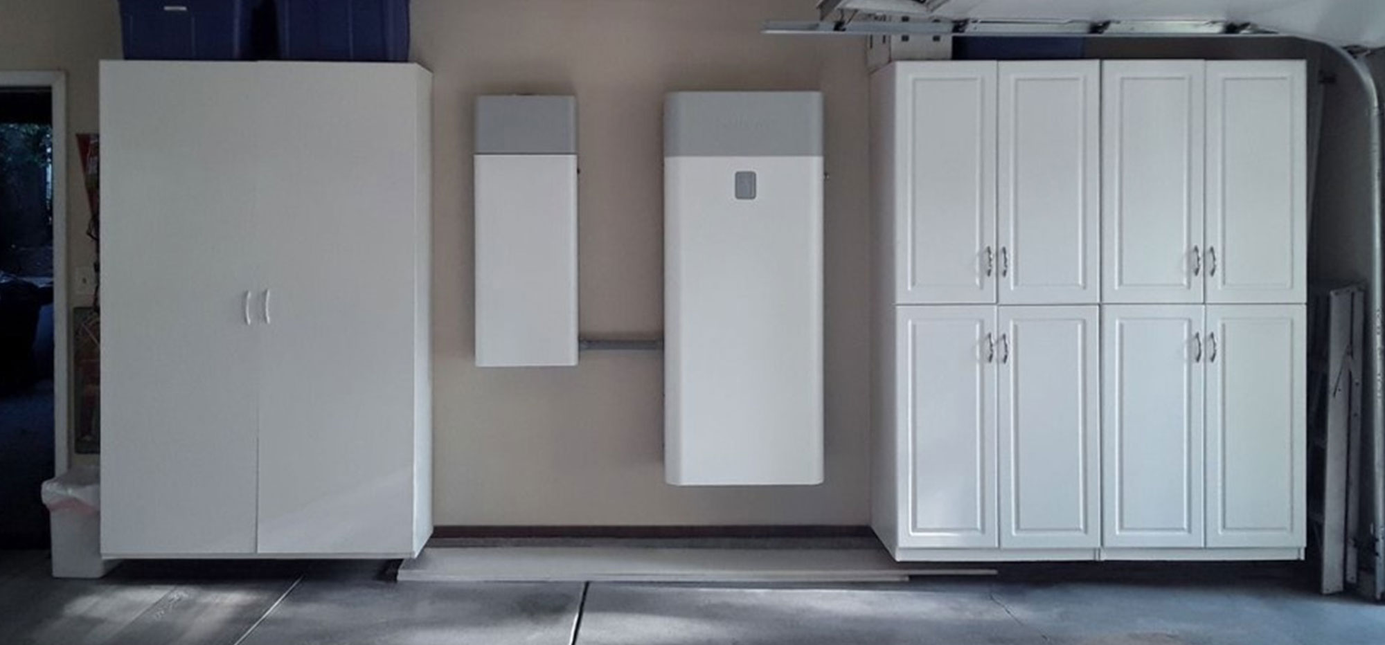 A garage interior with three large white storage units. On the left is a tall cabinet with two doors, in the center are two mounted rectangular power units, and on the right is a cabinet with four vertically aligned doors. A section of the garage floor is visible in front.