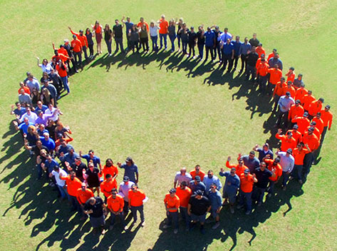 A large group of people standing in a circular formation on a grassy field. Many are wearing orange shirts, while others have dark or neutral-colored clothing. The photo is taken from above, showcasing the people in a shape resembling the outline of a large circle.