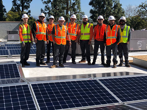 A group of nine construction workers wearing white helmets and bright orange safety vests stand together on a rooftop. They are surrounded by solar panels under a clear blue sky. Tall trees are visible in the background.