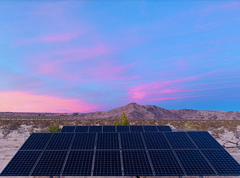 Solar panels are arranged in a flat array on a desert landscape with sparse vegetation. A mountain is visible in the background. The sky is vibrant, painted with shades of pink and blue, indicating either sunrise or sunset.