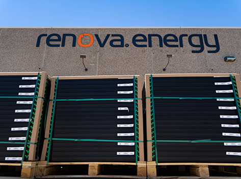 Stacks of solar panels are secured with green straps on wooden pallets outside a building. The building facade displays the logo renova.energy in bold letters. The sky above is clear and blue.