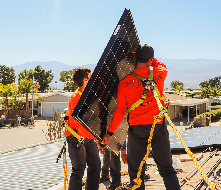 Three workers in orange shirts install a solar panel on a rooftop. They are wearing safety harnesses and are surrounded by suburban homes and trees, with mountains in the background. Its a sunny day, and the team appears focused on securing the panel in place.