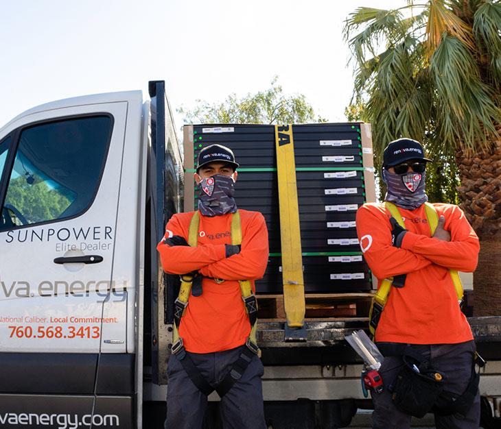 Two workers in orange shirts and sunglasses stand in front of a truck loaded with solar panels. Both are wearing face coverings and hard hats. The truck has company branding and text displaying Sunpower Elite Dealer and a phone number. Palm trees are visible in the background.