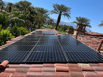 Solar panels are installed on a terracotta-tiled roof, surrounded by palm trees under a clear blue sky. The panels are positioned to maximize sunlight exposure. In the background, there are more trees and rooftops, with distant mountains visible on the horizon.