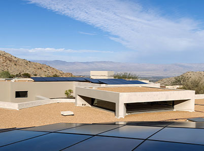 Modern, flat-roofed desert home with several solar panels on the roof. The house is set against a backdrop of arid hills and a bright blue sky. The landscape features sparse vegetation and expansive views of distant mountains.