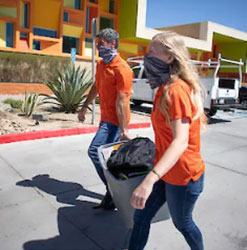 Two people wearing orange shirts and face coverings walk on a sunny sidewalk. They are holding a folded chair and a bag. The background features a building with colorful, geometric designs in shades of orange and yellow, with desert plants nearby.