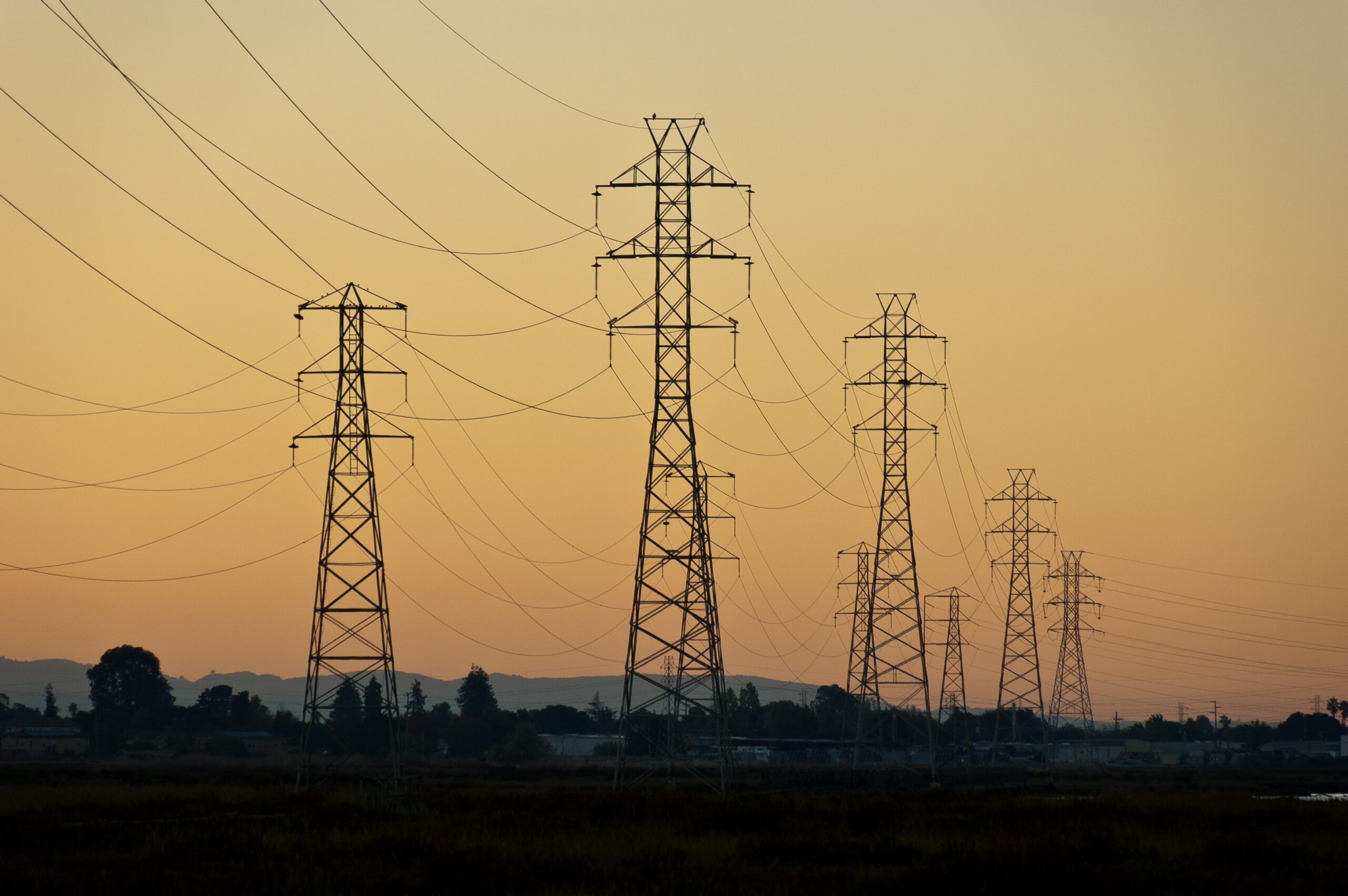 Power lines and lattice towers silhouetted against a golden sunset sky. The background features distant mountains and a clear gradient from orange to light yellow. The foreground shows dark trees and fields, enhancing the contrast with the colorful sky.