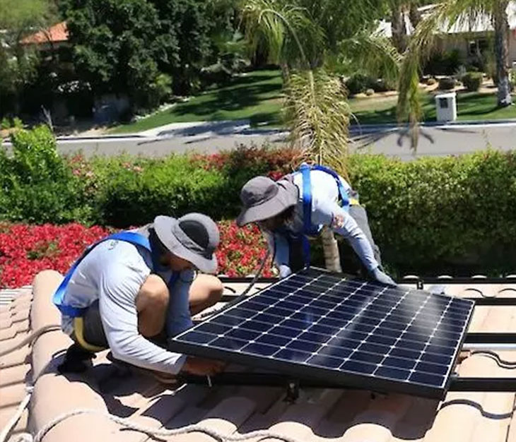 Two workers in blue uniforms and wide-brimmed hats are installing a solar panel on a red-tiled rooftop. They are crouched down, securing the panel in place. In the background, there are trees, a lawn, and a street with a mailbox visible. Sunny weather enhances the outdoor greenery.