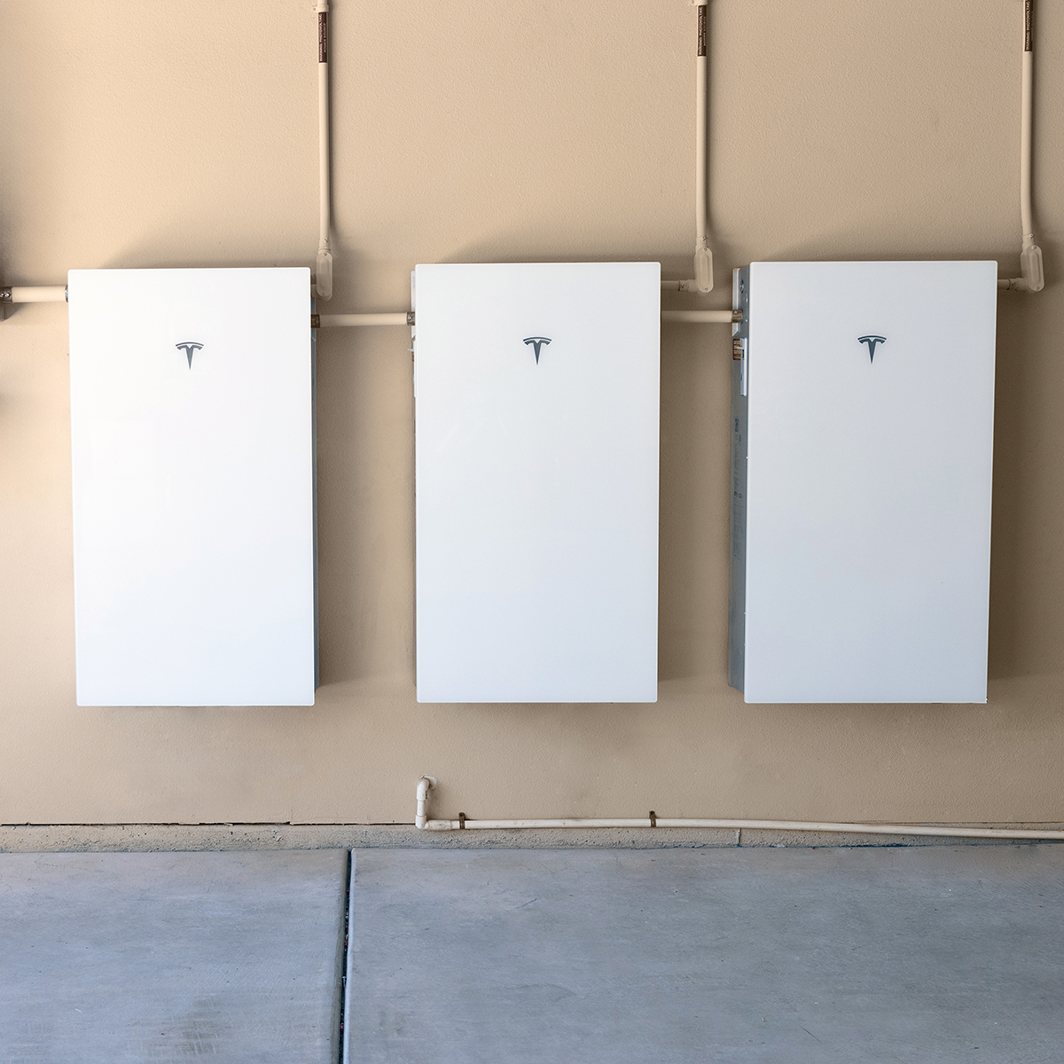 Tesla Collection 05 Three white Tesla Powerwall battery units are mounted side by side on a beige wall, installed by Palm Desert Solar Panel Installers, with visible wiring above and a concrete floor below.