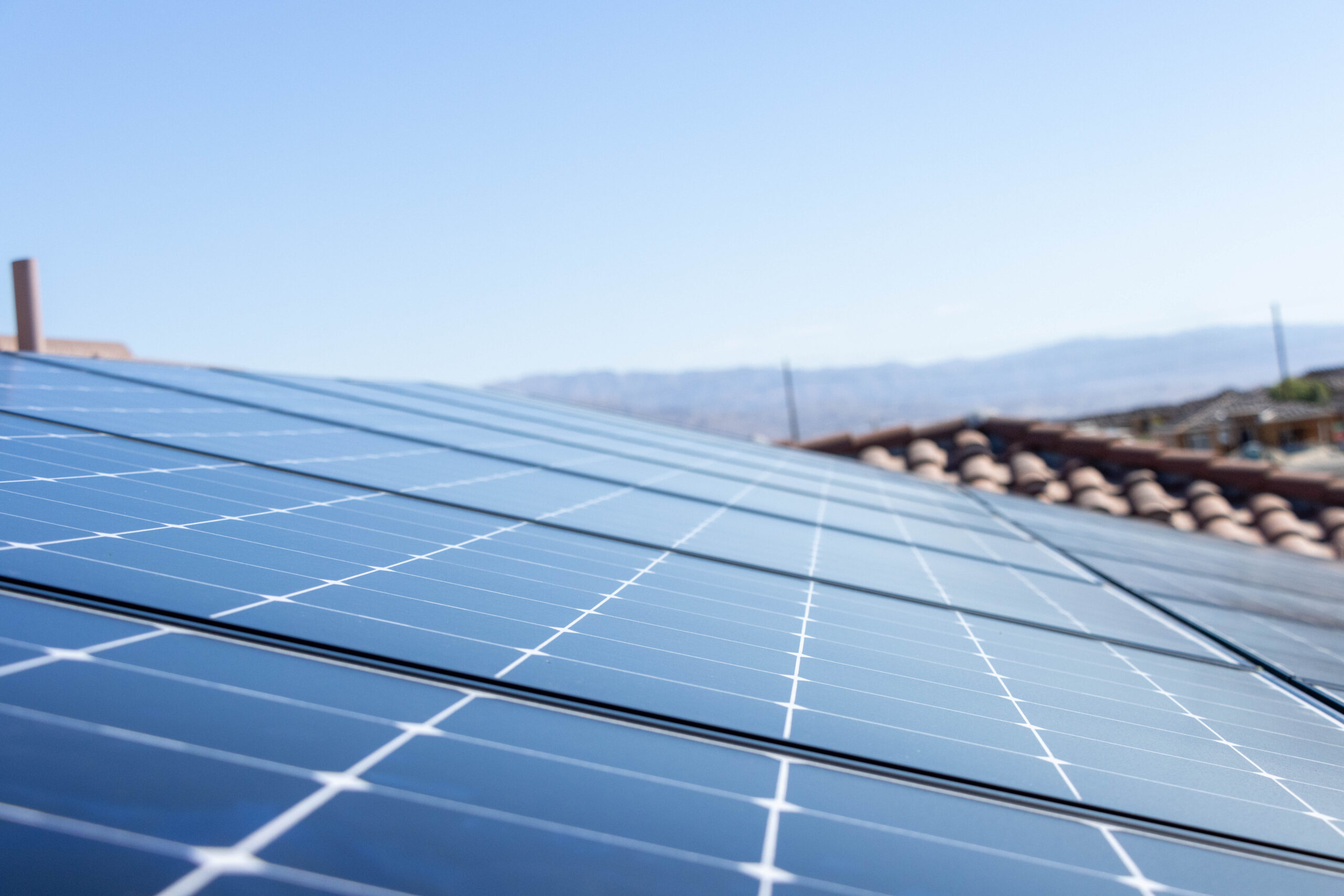 _DGT221019_6244 Close-up view of solar panels installed on a rooftop, with clear blue sky and distant mountains in the background. The panels reflect sunlight and are aligned in rows.
