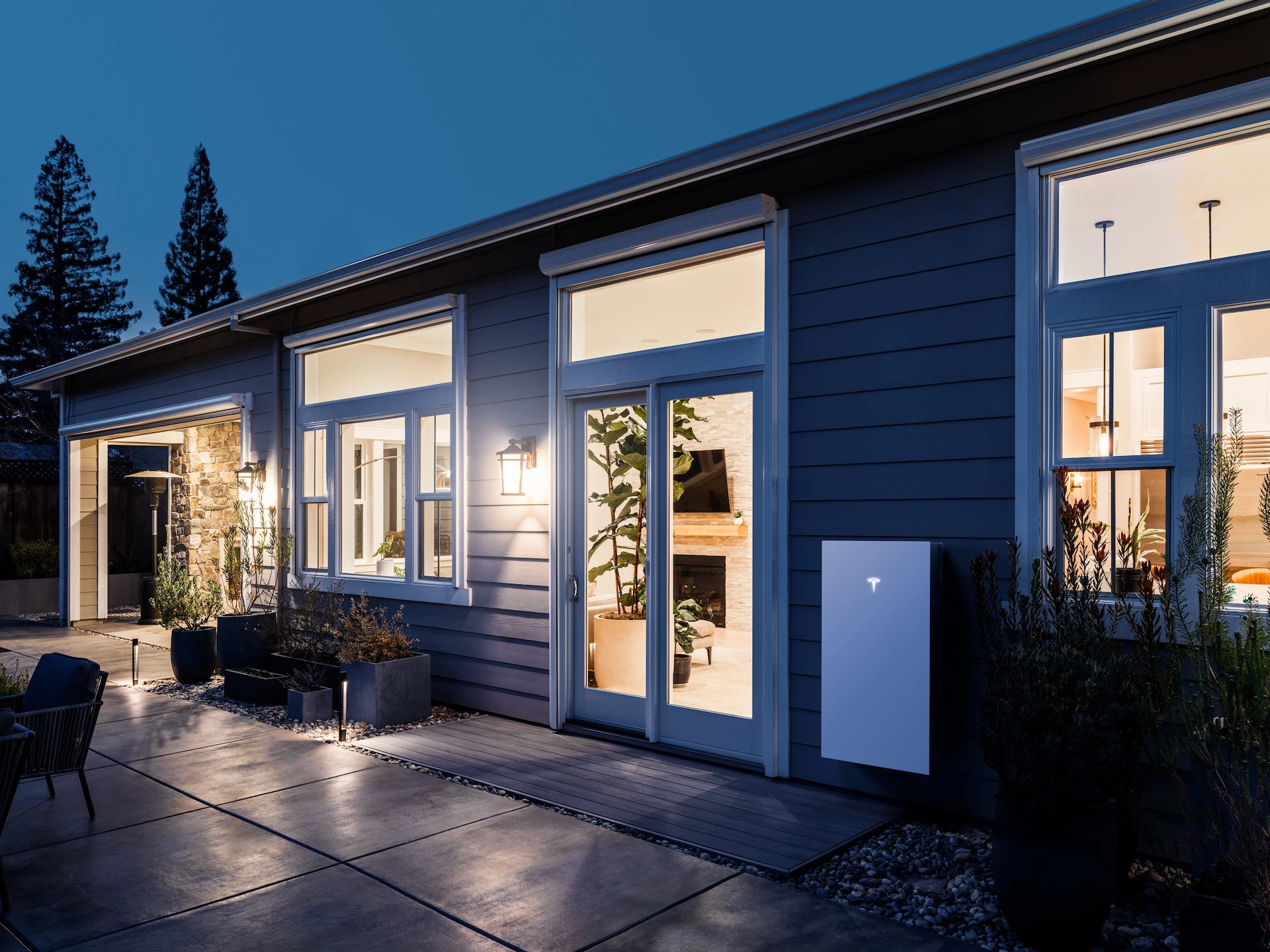Modern house exterior at dusk with large windows, lit interiors, potted plants, and a white Tesla Powerwall from Palm Springs Solar and Battery Systems mounted on the gray siding near the entrance.
