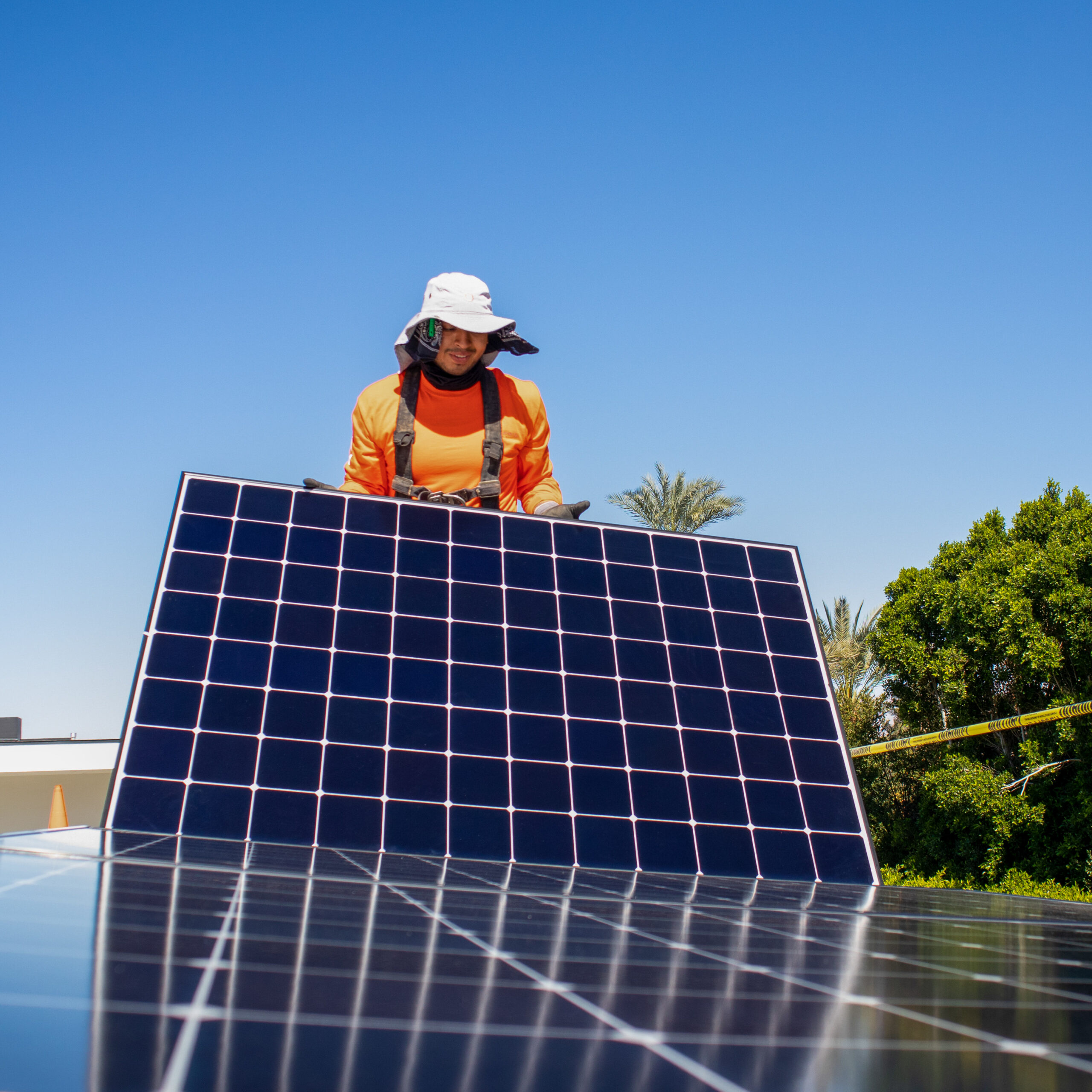 Sol_Panel1 A worker in an orange shirt and hat installs a solar panel outdoors on a sunny day, surrounded by trees and a clear blue sky—typical of the skilled Palm Desert Solar Panel Installers at work.