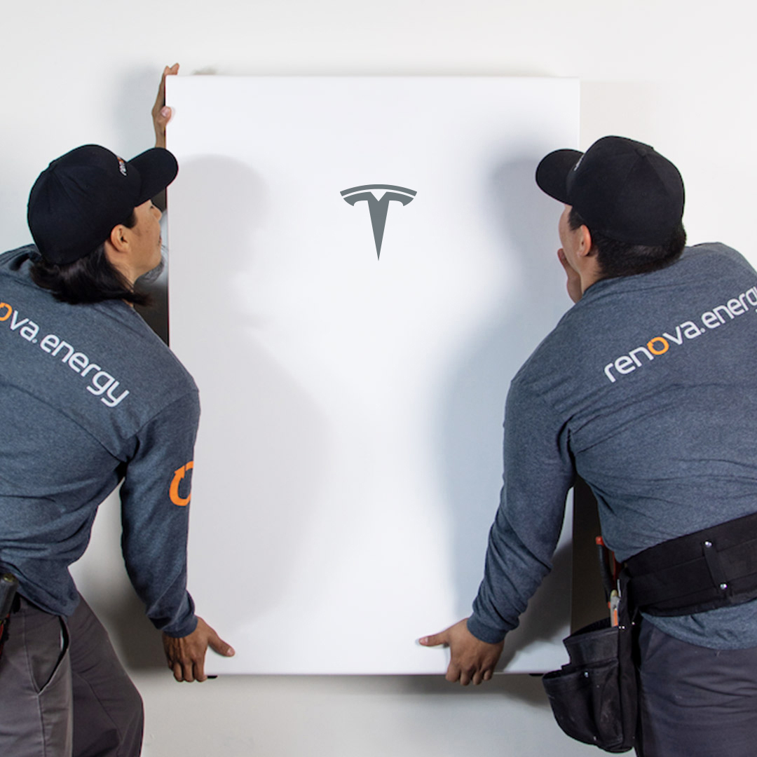 Two workers in Renova Energy uniforms are installing a white panel with the Tesla logo on it, setting up a reliable Palm Springs battery backup against the wall.