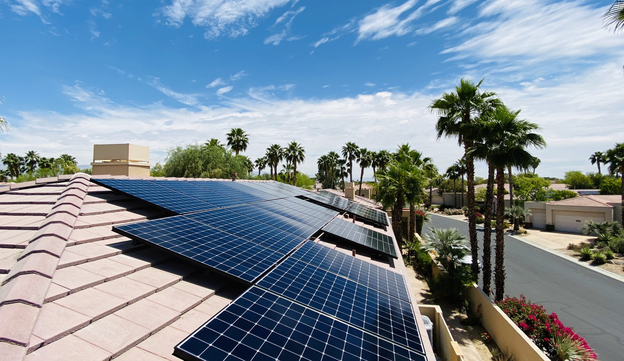 Rows of solar panels installed on a tiled rooftop in a sunny neighborhood, showcasing Palm Desert Solar Panel Installation with palm trees, houses, and a bright blue sky with scattered clouds in the background.
