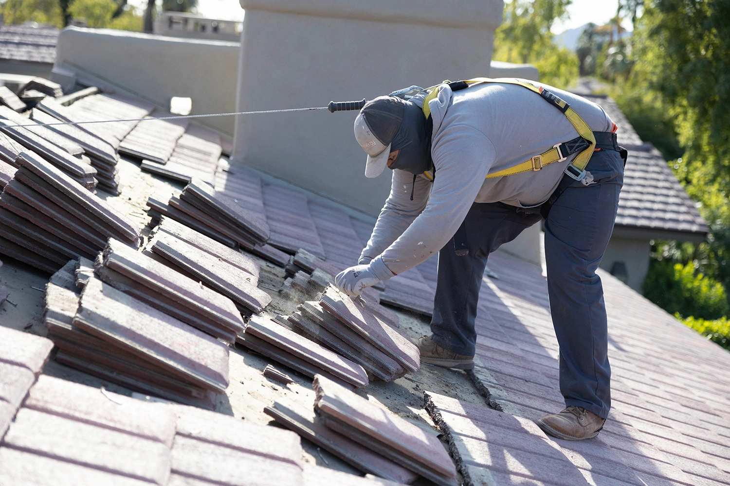 A construction worker wearing safety gear and a harness repairs roof tiles on a sloped rooftop in daylight, with some tiles removed and stacked. Indian Wells Solar Roofing stands out against trees and neighboring houses in the background.
