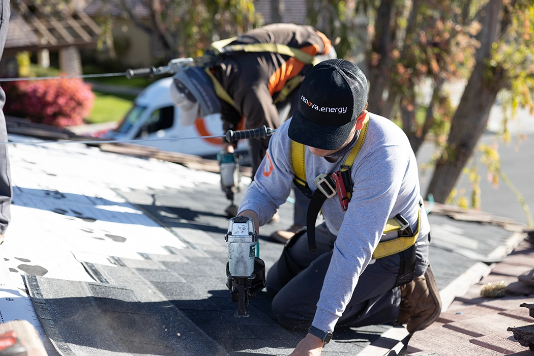 Two workers wearing safety harnesses install shingles on a rooftop for a Palm Springs Solar Roofing project. One uses a nail gun while the other works behind him. Its a sunny day with trees and a white van in the background.