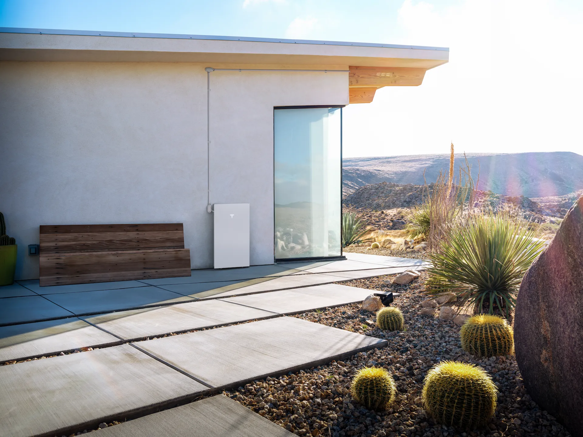 solar-battery-sytems-house-view A modern house with a large glass door, Tesla Powerwall battery by Palm Desert Solar and Battery Systems mounted outside, a wooden bench, and a desert landscape with cacti and rocky hills in the background.