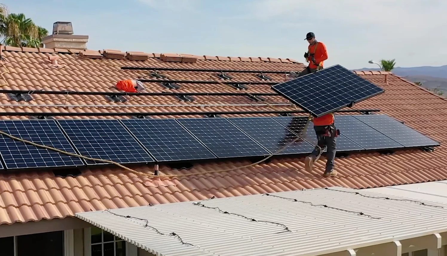 solar-roofing-renova Three workers in orange shirts install large solar panels on the red-tiled roof of a house under a clear sky. With Palm Desert Solar Roofing expertise, some panels are already in place while one worker carries another panel to be installed.