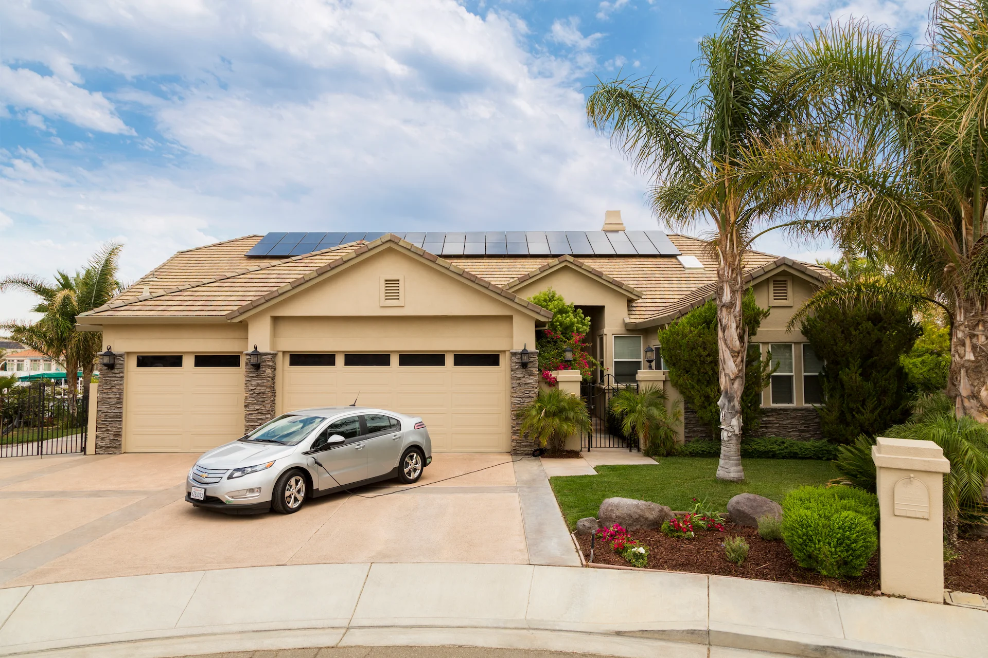 website_frontyard_with_panels_EV_original A modern beige house with solar panels by Indian Wells Solar and Battery Systems on the roof, a three-car garage, and a silver car in the driveway. Palm trees and a landscaped garden grace the front yard under a partly cloudy sky.