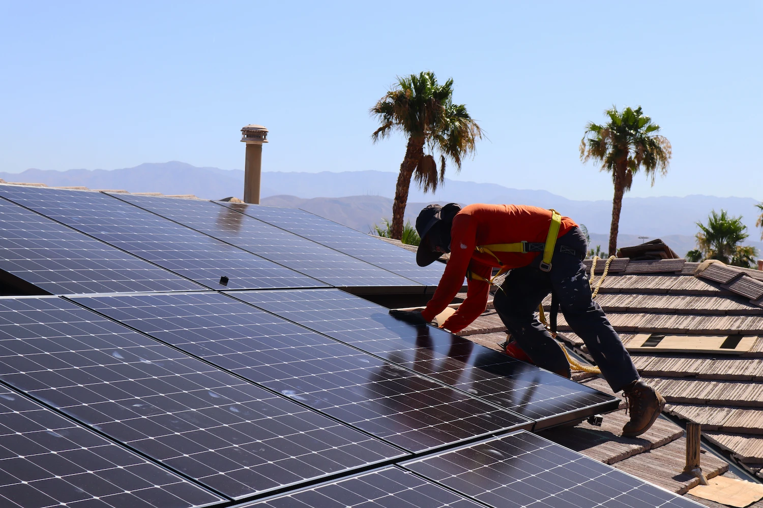 A worker wearing safety gear and a hat installs solar panels on a rooftop, showcasing Palm Springs Solar Maintenance with palm trees and mountains in the background under a clear blue sky.