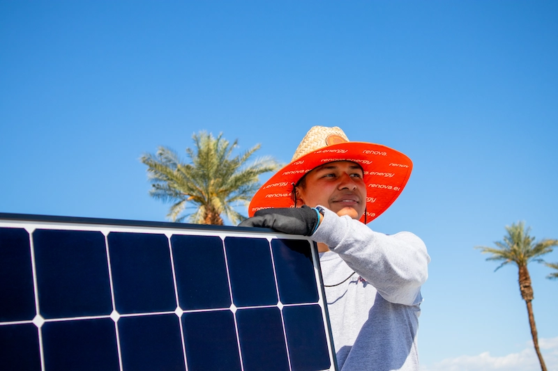 A person wearing an orange wide-brimmed hat and gloves installs a solar panel outdoors in Indian Wells, with palm trees and a clear blue sky in the background.