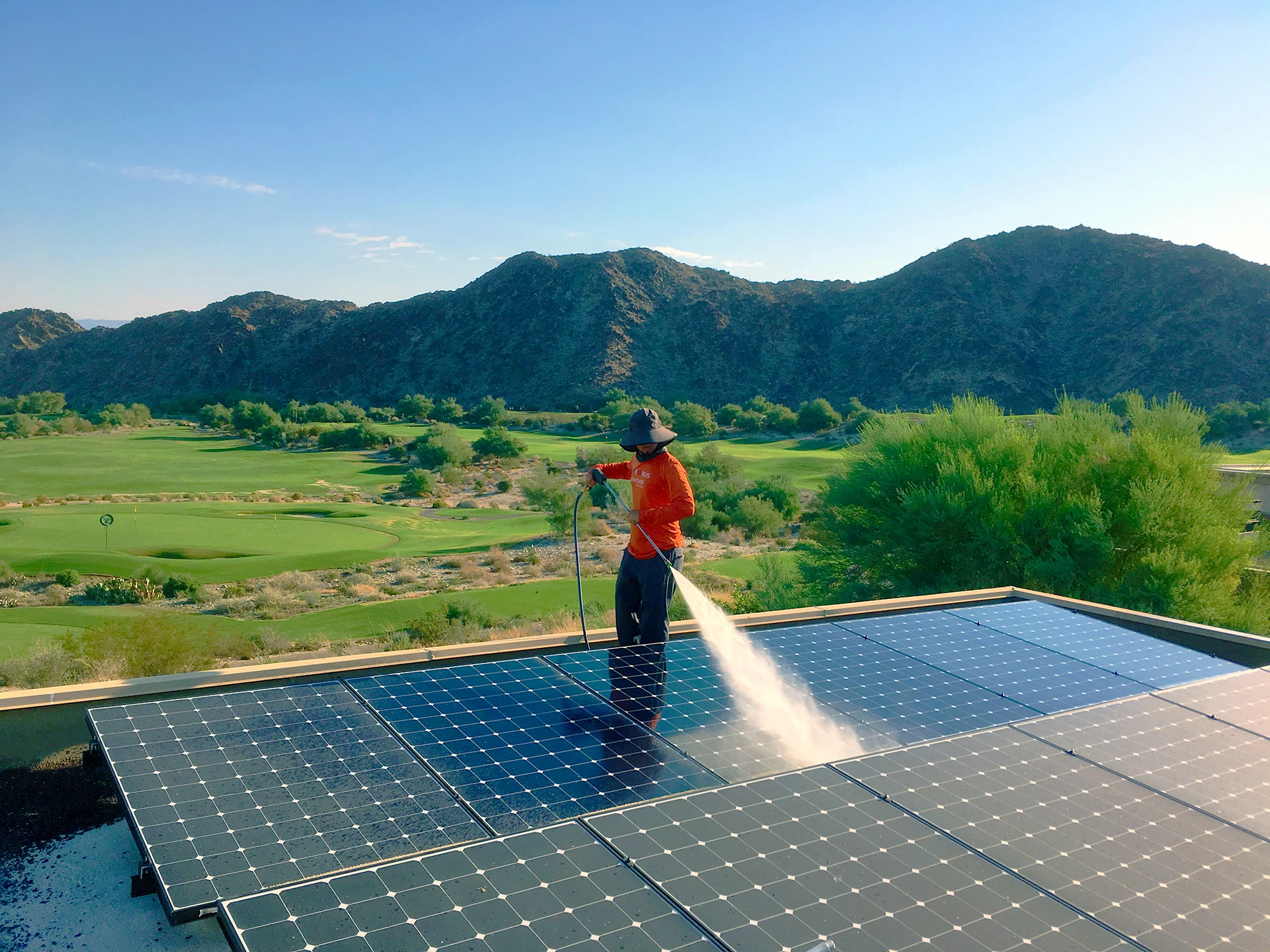 A person in a hat and orange shirt sprays water to clean solar panels on a rooftop—La Quinta Solar Maintenance ensures shining panels amid green landscape, trees, and mountains under a clear blue sky.