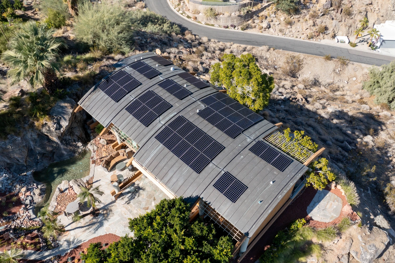 renova-business-rooftop-DGT221118_0315-1 Aerial view of a modern house with a curved roof covered in solar panels, surrounded by desert landscaping, trees, rocks, and a nearby winding road.