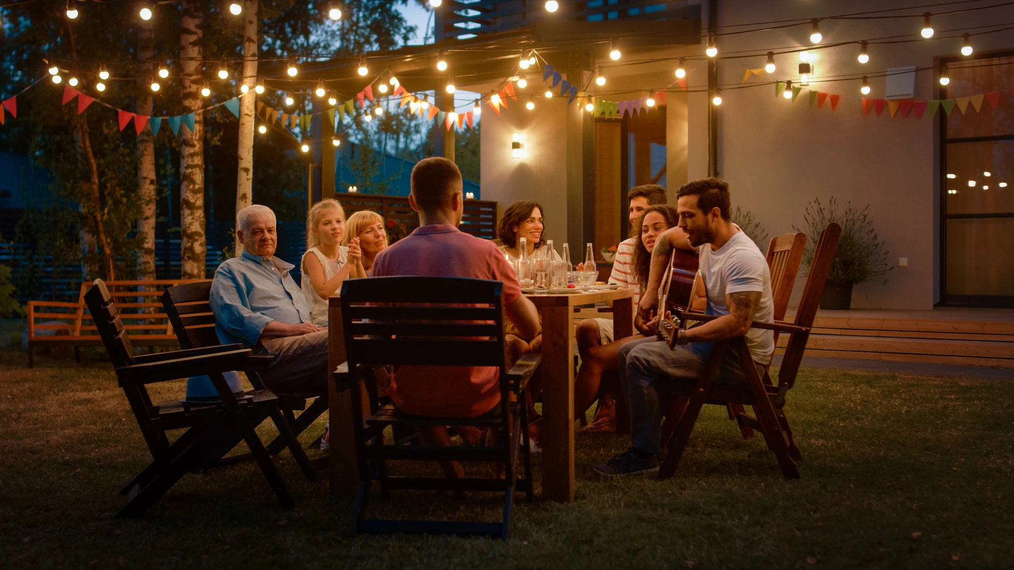 A group of people sit around a table outdoors in the evening, enjoying food and conversation under string lights powered by La Quinta Solar and Battery Systems. One person plays guitar, adding to the festive, warm scene filled with laughter and relaxed, happy expressions.