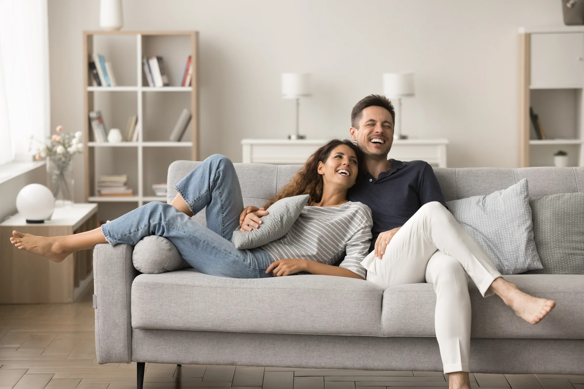 renova-happy-couple-on-couch-AS_1012139825 A smiling couple relaxes on a light gray sofa in a modern living room. The woman is lying with her legs across the mans lap, and both appear happy and comfortable together. Shelves and soft lighting are in the background.