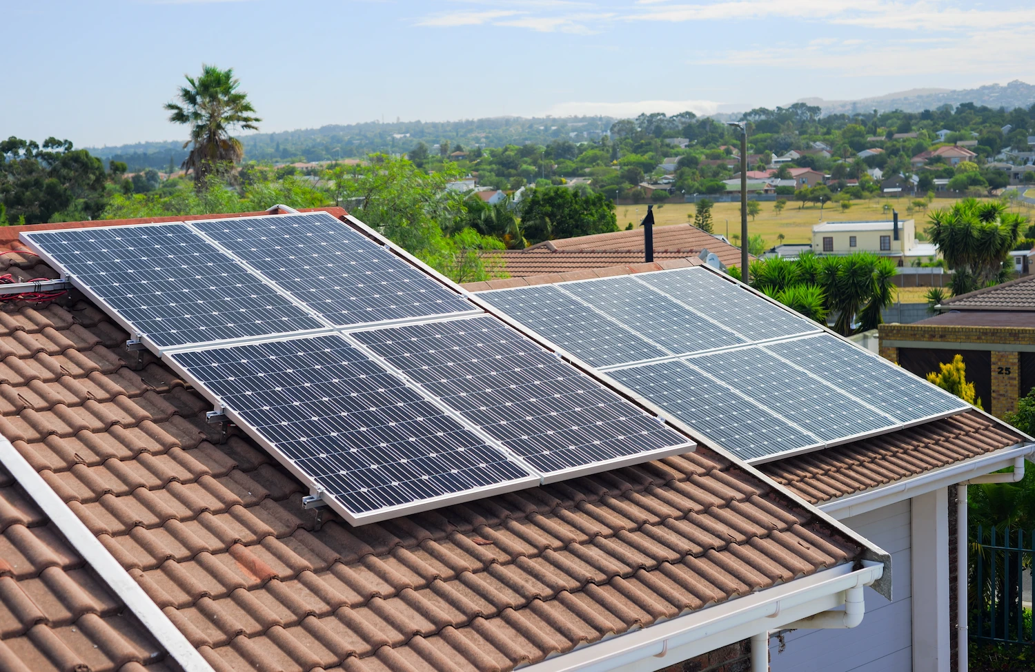 Solar panels installed on the tiled roof of a house in a suburban area with greenery, houses, and distant hills visible in the background under a partly cloudy sky.