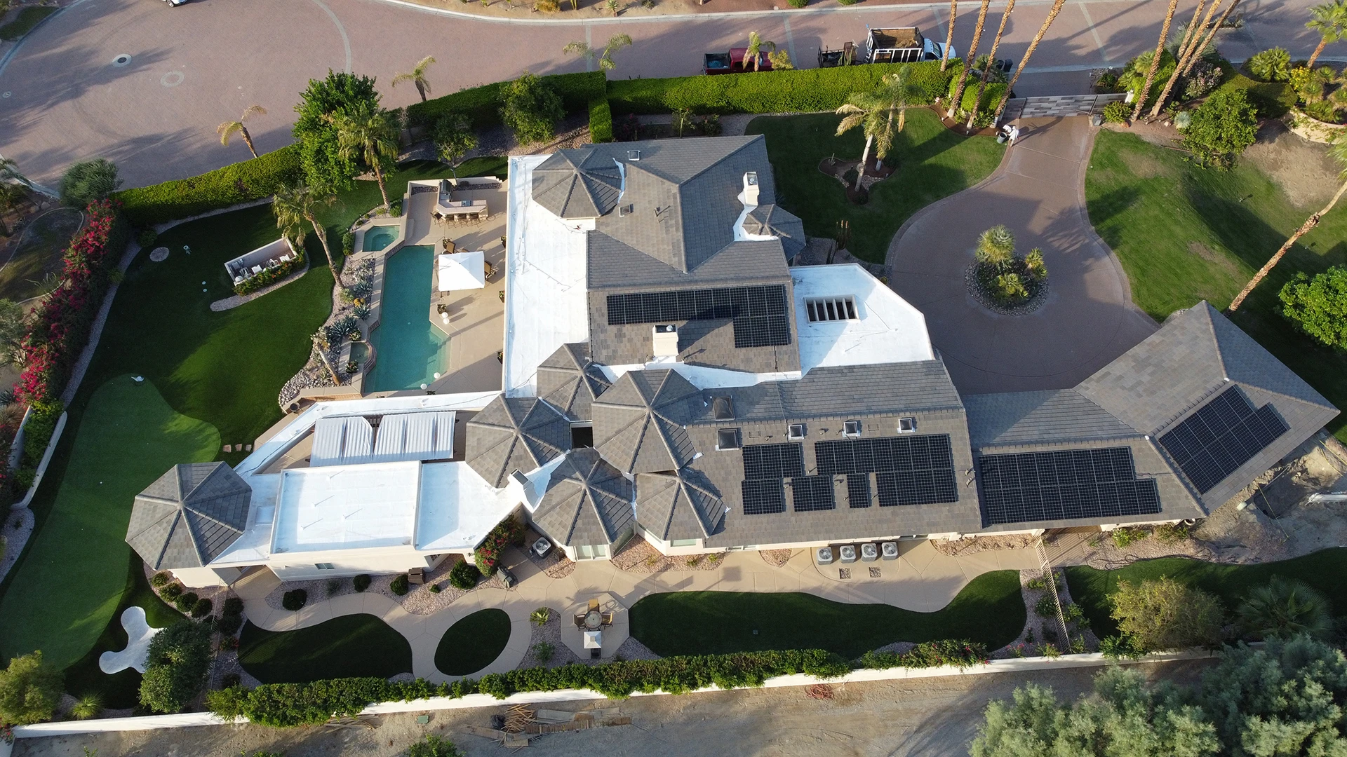re-house-roof-solar-panels-sm Aerial view of a large house with multiple gray roofs, a swimming pool, Cathedral City Solar and Battery Systems, landscaped gardens, palm trees, and curved driveways. Outdoor seating areas and green lawns surround the property.