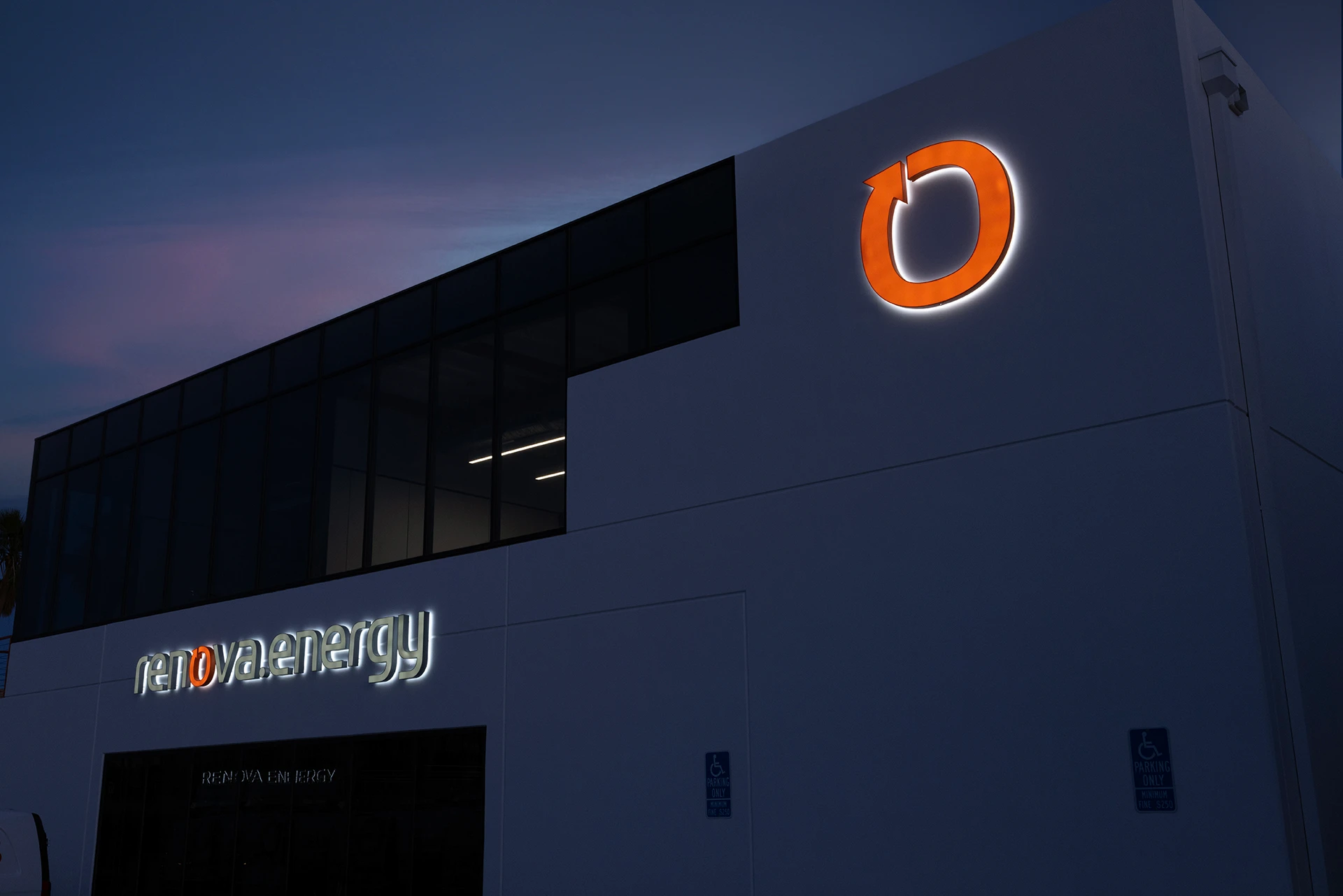 A modern white building at dusk with large glass windows and illuminated signs, including an orange circular logo and the text renova energy on the exterior wall, showcases Desert Hot Springs Solar Panel Installation expertise.